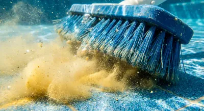Gros plan d'un balai de piscine soulevant un nuage de poussière jaune au fond de l'eau.